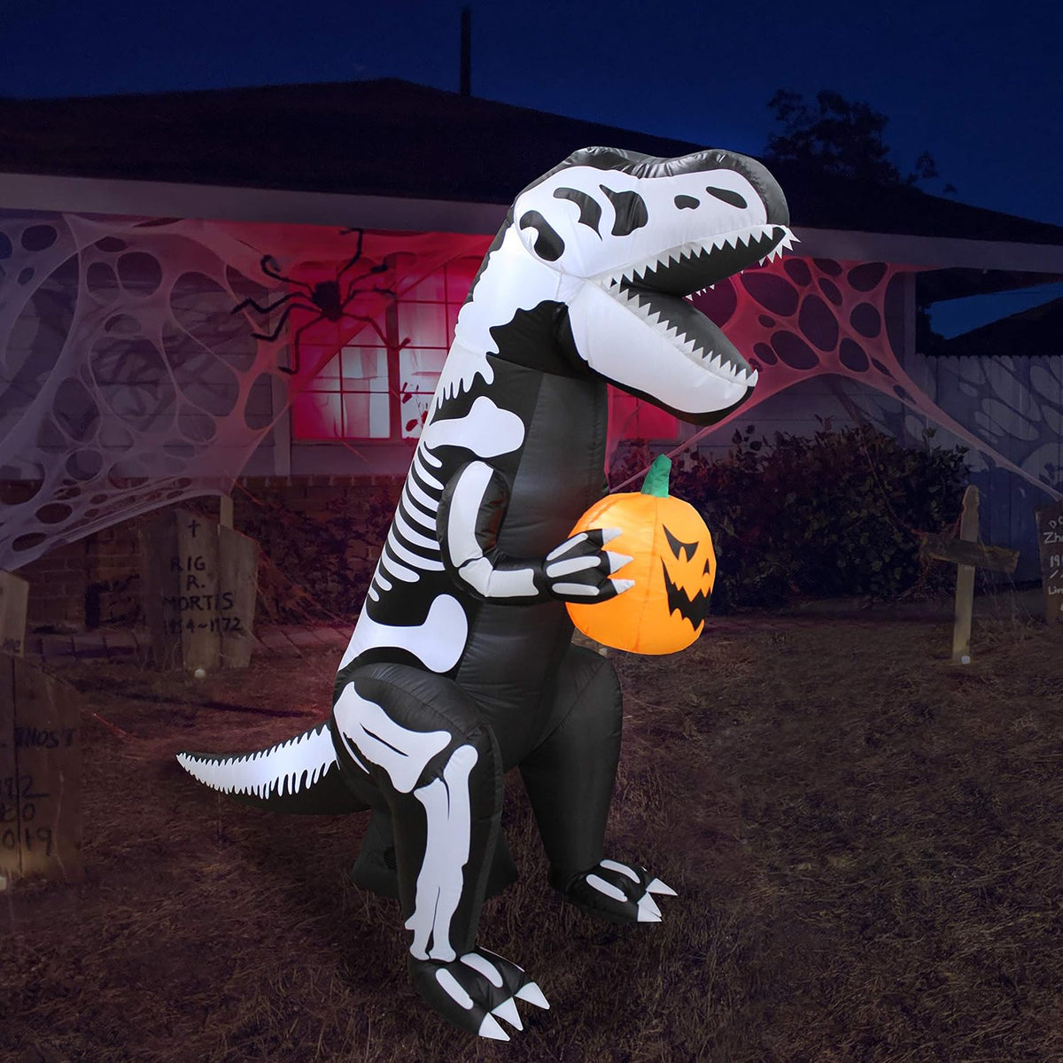 Inflatable dinosaur with a white skeleton print holding a jack-o'-lantern in a dark yard